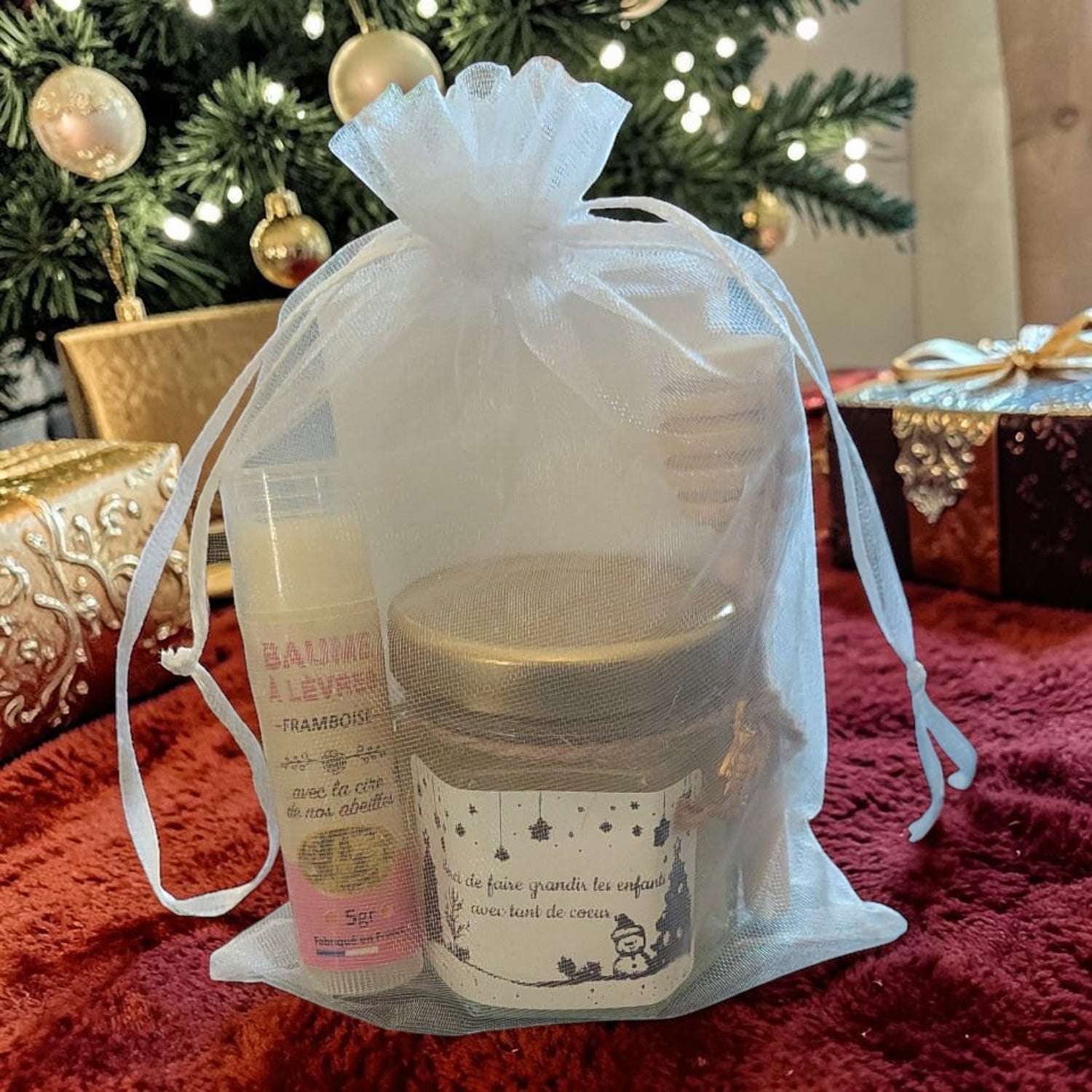 Cadeau de Noël personnalisé présenté sous le sapin avec mini pot de miel artisanal, baume à lèvres à la cire de nos abeilles, cuillère à miel en bois, breloque abeille et pochette organza prête à offrir.