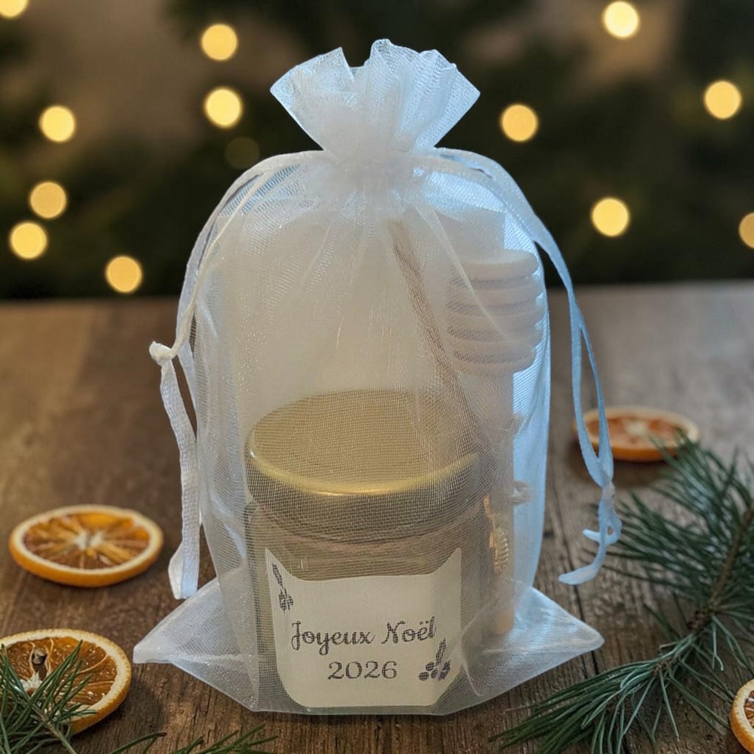 Mini pot de miel artisanal personnalisé avec étiquette Joyeux Noël 2026, cuillère à miel en bois, breloque abeille dorée et pochon en organza présenté dans une ambiance chaleureuse de Noël.