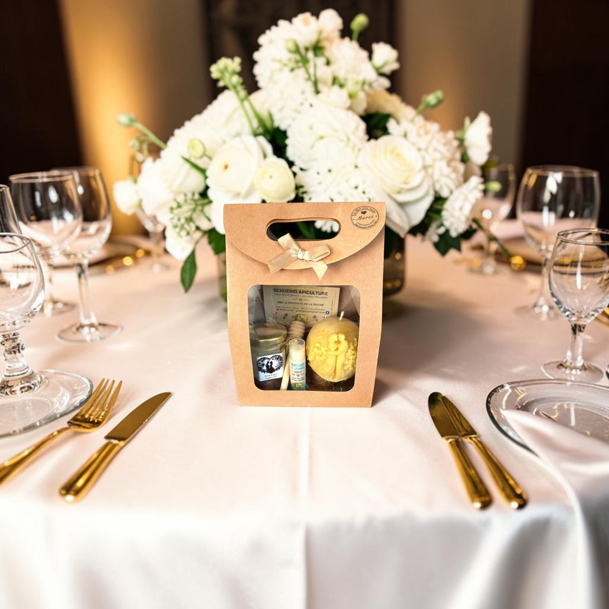 Coffret cadeau invité mariage personnalisé avec pot de miel, baume à lèvres artisanal et bougie en cire d'abeille, présenté sur une table de réception décorée de fleurs blanches.