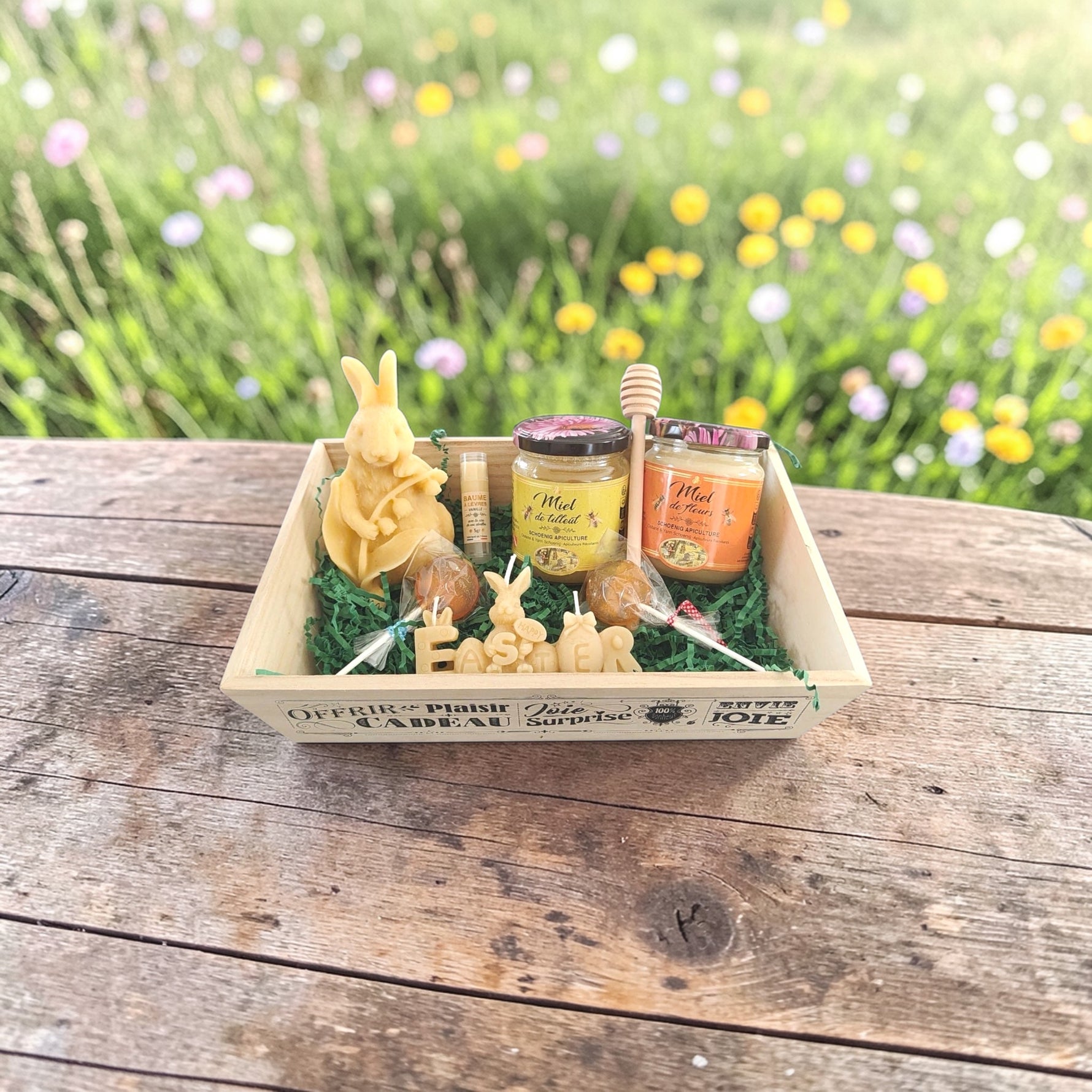 Coffret cadeau artisanal avec miel, bougies, sucettes, baume à lèvres et cuillère à miel en bois présenté sur une table en bois dans une prairie fleurie, idée cadeau naturelle et écologique.