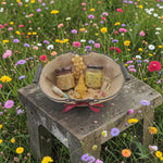 Coffret cadeau naturel avec miel, bougie en cire d’abeille et baume à lèvres posé sur une table en bois dans une prairie fleurie, ambiance champêtre et écologique.